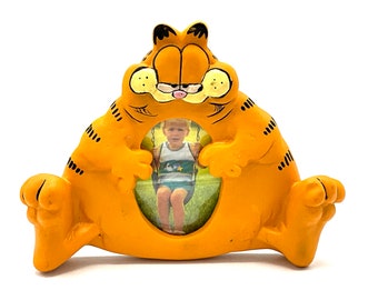 May include: Orange Garfield-shaped picture frame. The frame features the cartoon cat's face and body, with a photo opening in the center. The frame is made of a glossy material and has black stripes and details.