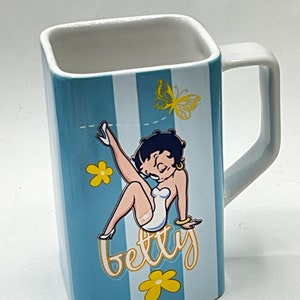 May include: A blue and white striped ceramic mug with a cartoon image of Betty Boop wearing a white dress and holding a yellow butterfly. The mug has the word "betty" written in yellow cursive letters.