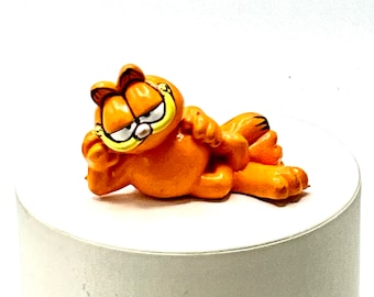 Puede incluir: Figurita de Garfield naranja tumbada con las patas cruzadas. La figurita está hecha de cerámica y tiene un estilo de dibujos animados.