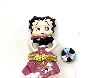 May include: A porcelain figurine of Betty Boop wearing a pink dress and black top, holding a record. She is standing on a wooden base with a small white dog beside her.