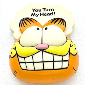 May include: Orange Garfield cartoon character figurine with a white speech bubble above his head that says "You Turn My Head!"