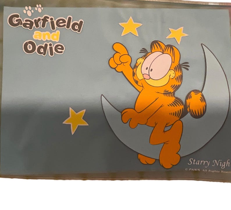 May include: A Garfield and Odie poster featuring Garfield sitting on a crescent moon, pointing upwards. The orange cat is surrounded by yellow stars against a blue background. The words "Garfield and Odie" are at the top.