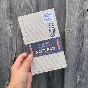 May include: A notepad with a blue cover and white lined paper. The cover has a sticker with a cartoon character and the text "It looks like you might forget something... Should you write it down?" The cover also has the text "mad love creative co NOTEPAD 4.25 x 7 in 25 sheets Notepad by Mad Love Creative Co."
