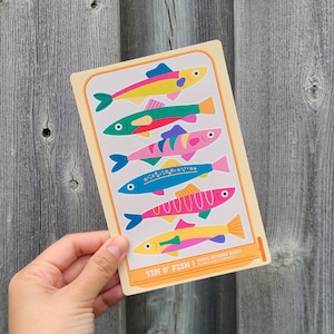 May include: A sticker sheet with six illustrated fish in bright colours. Each fish has a different design, with shades of yellow, pink, blue, and green. The text "TIN O' FISH" is visible at the bottom of the sheet.