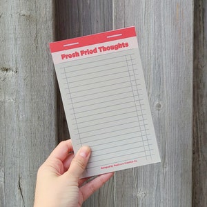 May include: A notepad with a red header and lined paper. The header says "Fresh Fried Thoughts" and is printed in a bold, white font. The notepad is held in a hand against a wood background.