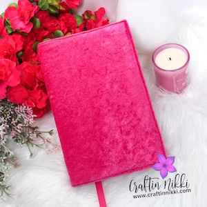May include: A hot pink velvet journal with a matching ribbon bookmark. The journal is rectangular and closed, with a soft, textured surface. A pink candle and red flowers are in the background. The website address "craftinnikki.com" is visible.