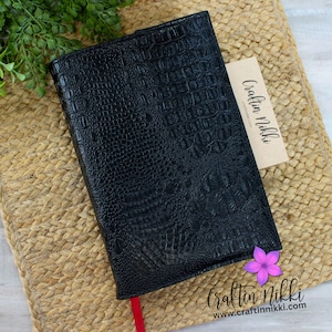 May include: Black faux leather book cover with a crocodile skin texture. A red ribbon bookmark is visible. A small tag with the text "Craftin Nikki" is tucked inside. A purple flower is placed near the bottom right corner.