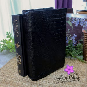 May include: A black leather-bound book with a crocodile-embossed pattern. The book is standing upright on a table next to a book titled "Onyx Storm" by Rebecca Yarros.