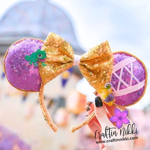 Peut inclure: Un serre-tête oreilles de souris avec un nœud doré à paillettes et des oreilles violettes à paillettes. Les oreilles sont décorées d'une grenouille verte et de rubans roses. Le bandeau est doré avec une bordure dorée à paillettes.