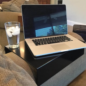May include: A black metal laptop tray sits on a brown couch. A laptop computer is on the tray, and a glass of water with ice cubes is on the tray to the left of the laptop.