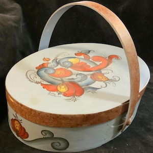 May include: A round, hand-painted wooden basket with a lid and handle. The lid features a floral design in red, blue, and yellow. The basket's body is light blue with a brown rim and handle. The artist's signature, "Betty Dahlquist", is visible on the lid.