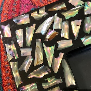 May include: Close-up of a black picture frame with iridescent shell fragments inlaid across the surface. The shell pieces reflect a range of colors, including white, gold, and green. The frame has a glossy black finish.