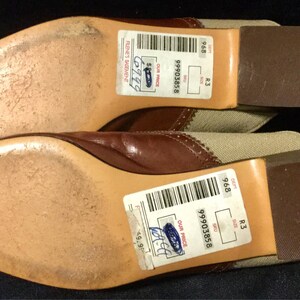 May include: A pair of brown leather shoes with beige canvas uppers. The shoes have a brown leather sole and a white stitching detail. The shoes have a price tag attached to them with the text "99903858" and "R3 9.68".