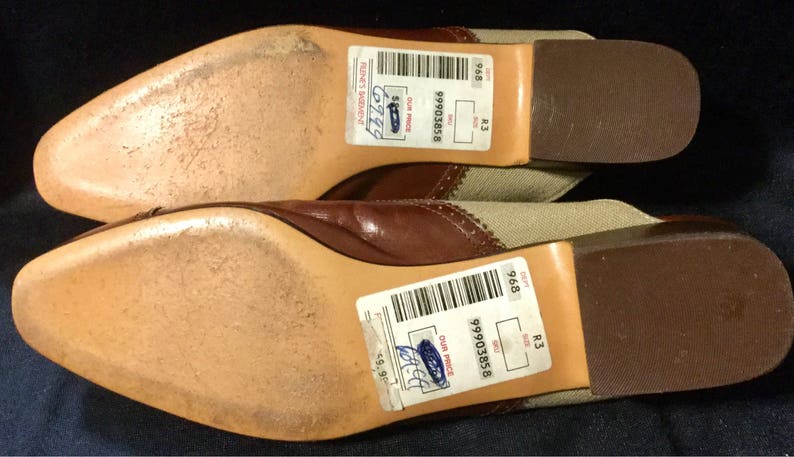 May include: A pair of brown leather shoes with beige canvas uppers. The shoes have a brown leather sole and a white stitching detail. The shoes have a price tag attached to them with the text "99903858" and "R3 9.68".