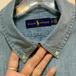 May include: Light blue denim shirt with a button-down collar. The shirt features a navy blue label with the text "RALPH LAUREN" and a yellow polo player logo. The shirt is size XL.