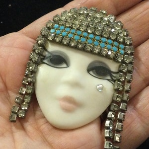 May include: A vintage brooch in the shape of a stylized face. The face is white with painted features, including large eyes and a small mouth. The headpiece and hair are adorned with clear rhinestones and turquoise accents.