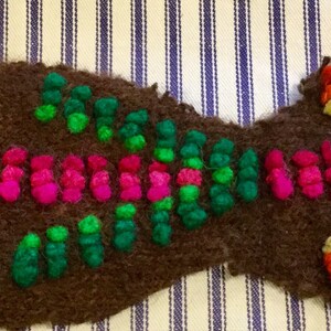 K&ouml;nnte beinhalten: Ein braunes Stoffst&uuml;ck mit bunter Stickerei. Die Stickerei zeigt ein Blumenmuster mit rosa, gr&uuml;nem, blauem und wei&szlig;em Garn. Die Blume ist von einem Muster aus rosa, gr&uuml;nem und orangefarbenem Garn umgeben.