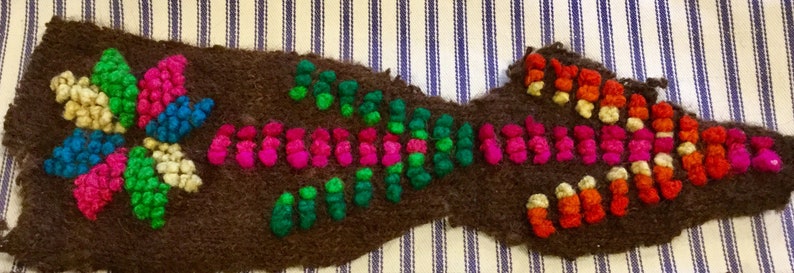K&ouml;nnte beinhalten: Ein braunes Stoffst&uuml;ck mit bunter Stickerei. Die Stickerei zeigt ein Blumenmuster mit rosa, gr&uuml;nem, blauem und wei&szlig;em Garn. Die Blume ist von einem Muster aus rosa, gr&uuml;nem und orangefarbenem Garn umgeben.