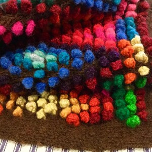 K&ouml;nnte beinhalten: Ein bunter, handgewebter Teppich mit braunem Hintergrund und einer Vielzahl von bunten Garnpompons. Der Teppich besteht aus verschiedenen Quadraten mit unterschiedlichen Farben und Mustern.