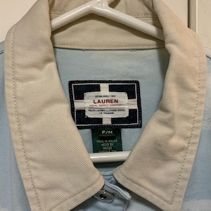 May include: Light blue denim jacket with a cream-colored collar and the "LAUREN NAVAL SUPPLY COMPANY" label. The jacket is hanging on a white hanger. The label also includes the text "MADE IN MACAU".