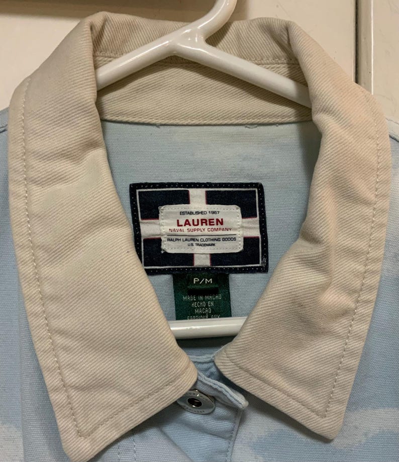 May include: Light blue denim jacket with a cream-colored collar and the "LAUREN NAVAL SUPPLY COMPANY" label. The jacket is hanging on a white hanger. The label also includes the text "MADE IN MACAU".