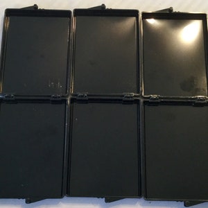 May include: Six black rectangular hinged containers are open, revealing empty compartments. The containers are arranged in two rows of three. Each container has a shiny, reflective interior and a matte black exterior.