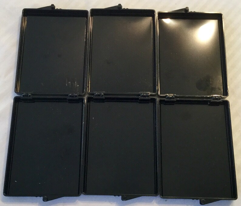 May include: Six black rectangular hinged containers are open, revealing empty compartments. The containers are arranged in two rows of three. Each container has a shiny, reflective interior and a matte black exterior.
