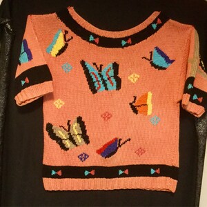 K&ouml;nnte beinhalten: Ein rosa gestrickter Pullover mit kurzen &Auml;rmeln und einem Rundhalsausschnitt. Der Pullover hat ein farbenfrohes Schmetterlingsdesign mit schwarzen, blauen, gelben, roten und gr&uuml;nen Akzenten.