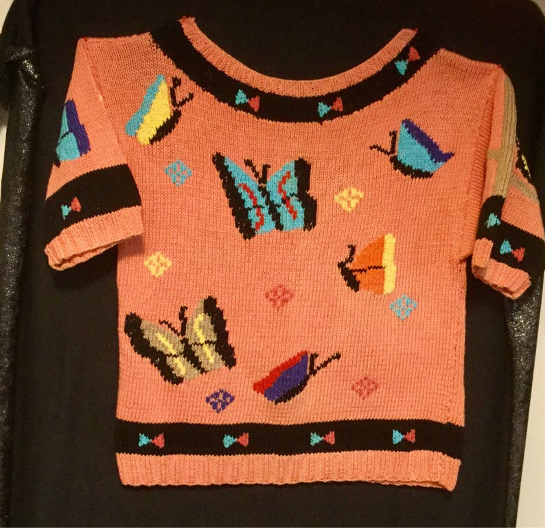 K&ouml;nnte beinhalten: Ein rosa gestrickter Pullover mit kurzen &Auml;rmeln und einem Rundhalsausschnitt. Der Pullover hat ein farbenfrohes Schmetterlingsdesign mit schwarzen, blauen, gelben, roten und gr&uuml;nen Akzenten.