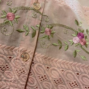 May include: Close-up of a pale pink garment with floral embroidery. The embroidery features pink roses and green leaves with gold-coloured stems. The garment has a button-up front with clear buttons and a decorative eyelet lace trim.