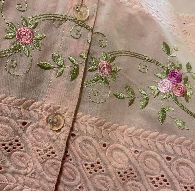 May include: Close-up of a pale pink garment with floral embroidery. The embroidery features pink roses and green leaves with gold-coloured stems. The garment has a button-up front with clear buttons and a decorative eyelet lace trim.