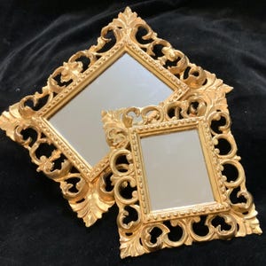 May include: Two ornate, gold-colored mirrors with square frames. The frames feature intricate, swirling designs. The mirrors are of different sizes, with the larger one partially obscuring the smaller one. The background is a dark, textured fabric.