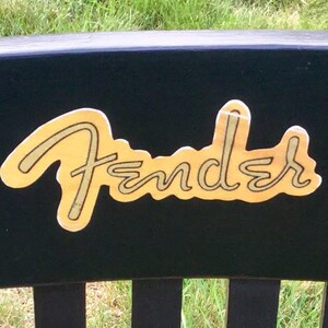Op de afbeelding: Zwarte houten stoel met een gouden Fender-logo en zilveren stemknoppen.