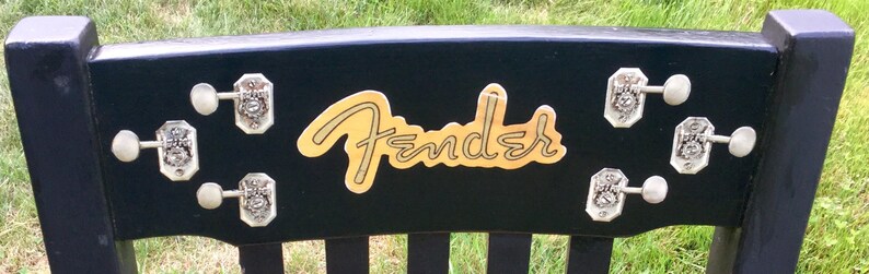 Op de afbeelding: Zwarte houten stoel met een gouden Fender-logo en zilveren stemknoppen.