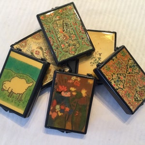 May include: A collection of small, rectangular boxes with black frames. The boxes feature various decorative designs, including floral patterns, a sheep in a field, and ornate gold and green embellishments. The boxes are arranged in a slightly overlapping manner.