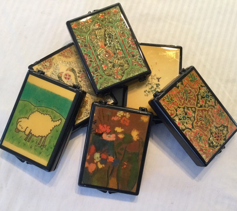 May include: A collection of small, rectangular boxes with black frames. The boxes feature various decorative designs, including floral patterns, a sheep in a field, and ornate gold and green embellishments. The boxes are arranged in a slightly overlapping manner.
