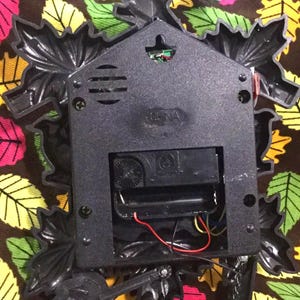 Op de afbeelding: De achterkant van een zwarte koekoeksklok met een vogel- en bladontwerp. De klok heeft een batterijvak en een kleine luidspreker. De klok is gemaakt van plastic en heeft een zwarte afwerking.