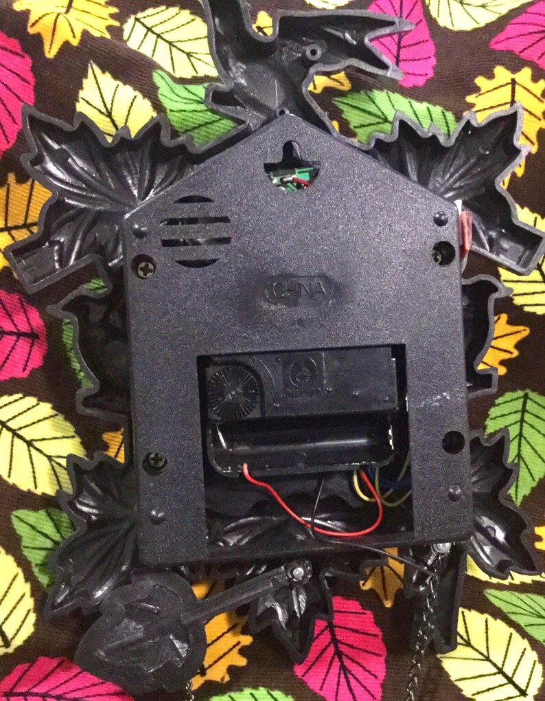 Op de afbeelding: De achterkant van een zwarte koekoeksklok met een vogel- en bladontwerp. De klok heeft een batterijvak en een kleine luidspreker. De klok is gemaakt van plastic en heeft een zwarte afwerking.