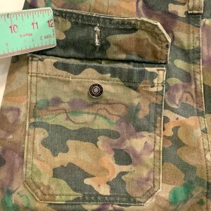 May include: Close-up of a camouflage-patterned pocket on denim pants. The pocket features a metal button and is accented with stitching. A ruler shows a measurement of approximately 10 to 12 inches.
