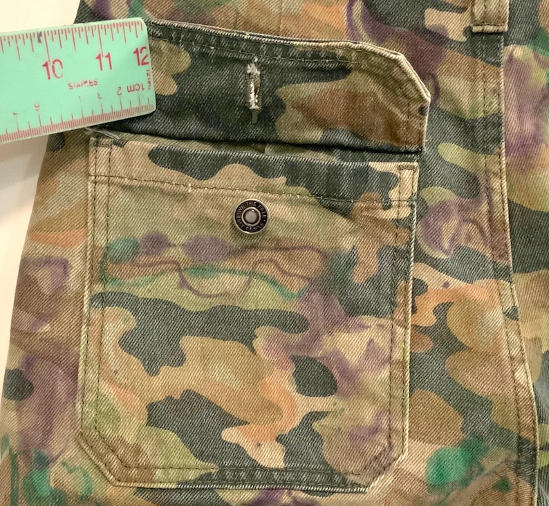 May include: Close-up of a camouflage-patterned pocket on denim pants. The pocket features a metal button and is accented with stitching. A ruler shows a measurement of approximately 10 to 12 inches.