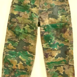 May include: Camouflage-patterned pants in shades of green, brown, and tan. The pants have a button and zipper closure, front pockets, and belt loops. The fabric appears to be a durable cotton blend. The camouflage design is a mix of organic shapes.