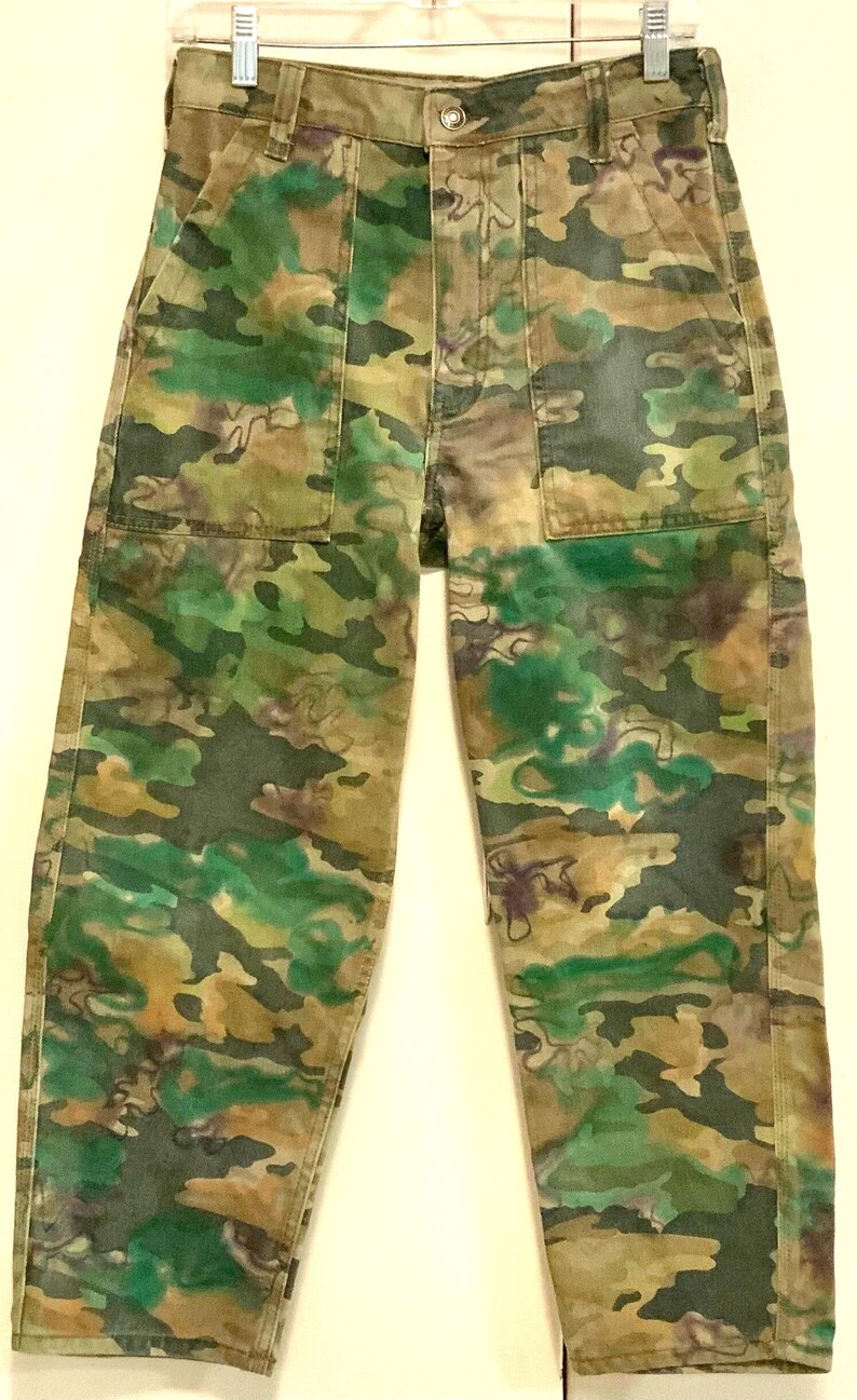 May include: Camouflage-patterned pants in shades of green, brown, and tan. The pants have a button and zipper closure, front pockets, and belt loops. The fabric appears to be a durable cotton blend. The camouflage design is a mix of organic shapes.