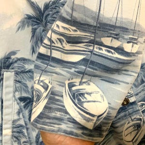 May include: Light blue button-up shirt with a nautical print featuring sailboats and palm trees. The fabric has a detailed pattern of white boats on a blue sea, with palm trees in the background. The shirt's sleeve is visible, showing the print.