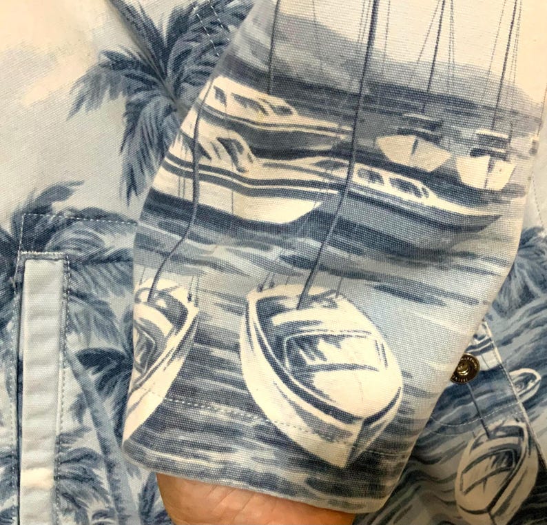 May include: Light blue button-up shirt with a nautical print featuring sailboats and palm trees. The fabric has a detailed pattern of white boats on a blue sea, with palm trees in the background. The shirt's sleeve is visible, showing the print.