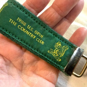 May include: Green fabric keychain with gold text that reads "1988 U.S. OPEN THE COUNTRY CLUB" and a gold squirrel graphic. The keychain has a metal clasp.
