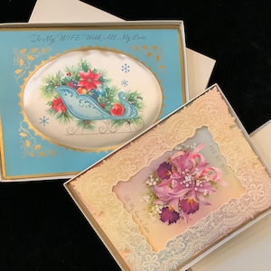 May include: Two vintage greeting cards with lace trim. The first card is blue with a gold trim and features a sleigh with Christmas decorations. The second card is cream with a pink ribbon and features a bouquet of purple flowers. The text on the cards reads "To My Wife With All My Love" and "For My Wife All My Love at Easter".