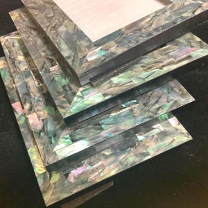 May include: A stack of rectangular picture frames with iridescent, mosaic-like inlays. The frames have a dark, possibly black, border and a light-colored backing. The mosaic pieces reflect shades of green, purple, and silver.