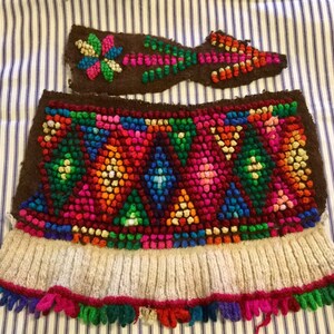 K&ouml;nnte beinhalten: Ein braunes gestricktes Gewebe mit einem bunten geometrischen Muster aus kleinen Wollkugeln. Das Gewebe hat einen wei&szlig;en gestrickten Rand und einen Fransen aus buntem Garn.