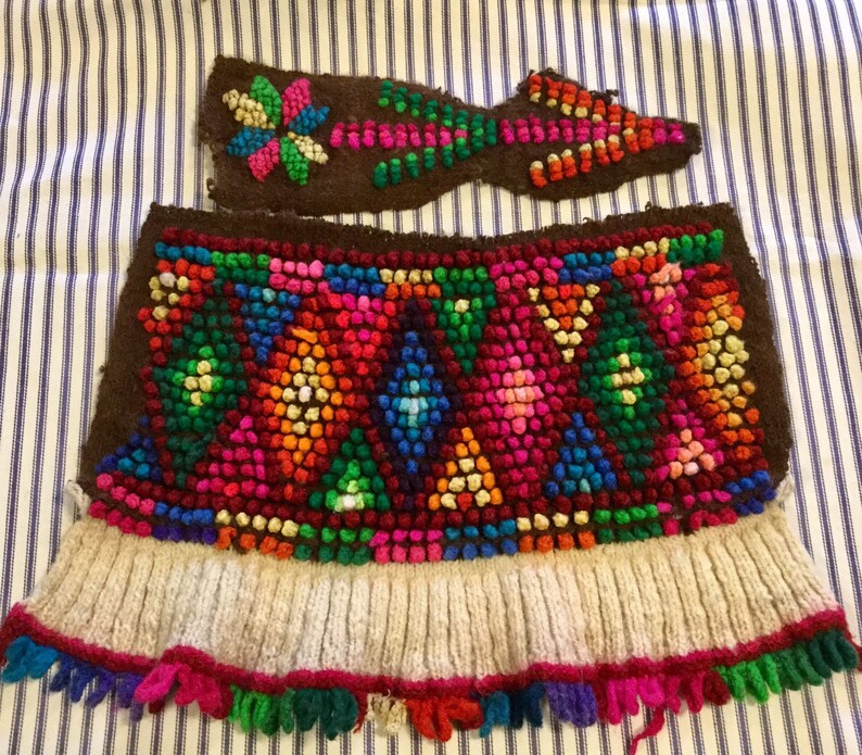 K&ouml;nnte beinhalten: Ein braunes gestricktes Gewebe mit einem bunten geometrischen Muster aus kleinen Wollkugeln. Das Gewebe hat einen wei&szlig;en gestrickten Rand und einen Fransen aus buntem Garn.