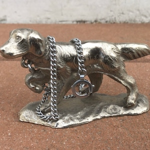 May include: A silver metal sculpture of a dog wearing a chain necklace. The dog is standing on a rock-like base.